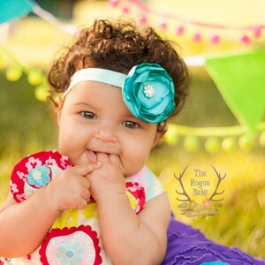 May include: A baby girl with curly brown hair wearing a turquoise flower headband with a rhinestone center. She is wearing a colorful floral dress and is sitting on a purple blanket in a grassy area.