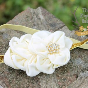 May include: A white satin flower headband with a gold glitter elastic band and a rhinestone centre. The headband is on a brown textured surface.