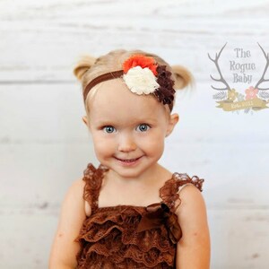 May include: A young girl with blonde hair wearing a brown lace dress and a headband with a brown, white, and orange flower. The headband is on a white background. The girl is smiling.