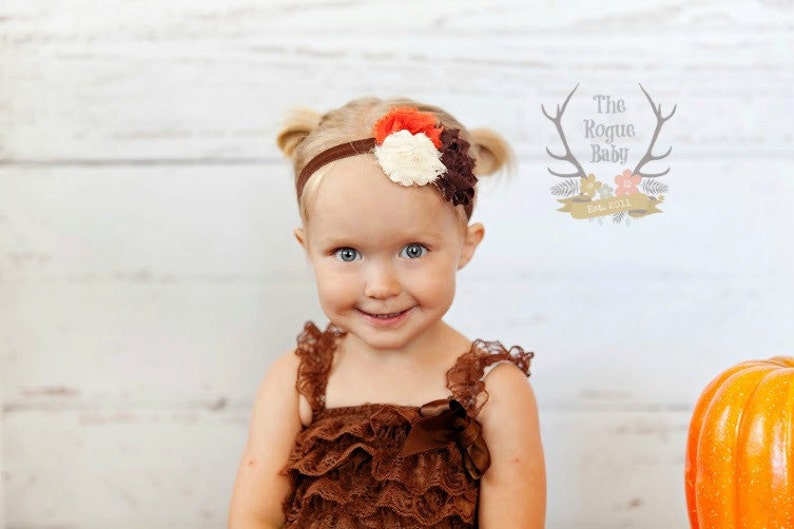 May include: A young girl with blonde hair wearing a brown lace dress and a headband with a brown, white, and orange flower. The headband is on a white background. The girl is smiling.