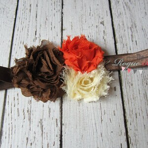 May include: A brown, orange, and cream fabric flower headband with a brown elastic band. The headband has the text "The Rogue Baby" printed on it.
