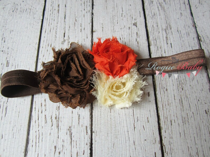 May include: A brown, orange, and cream fabric flower headband with a brown elastic band. The headband has the text "The Rogue Baby" printed on it.