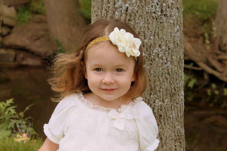 May include: A young girl with long brown hair wearing a white dress with a lace trim and a gold headband with a white flower accent.