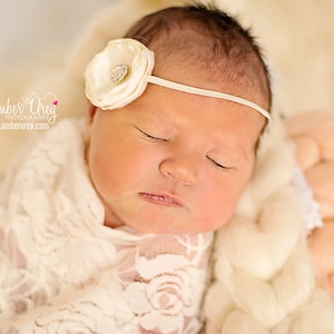 May include: A newborn baby girl sleeping peacefully in a white lace blanket. She is wearing a white headband with a white flower and a rhinestone.