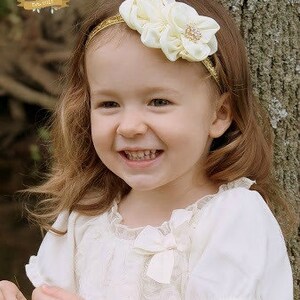May include: A young girl with long brown hair is wearing a white dress with a lace overlay and a gold headband with a white flower accent. The headband has a small rhinestone in the centre of the flower.