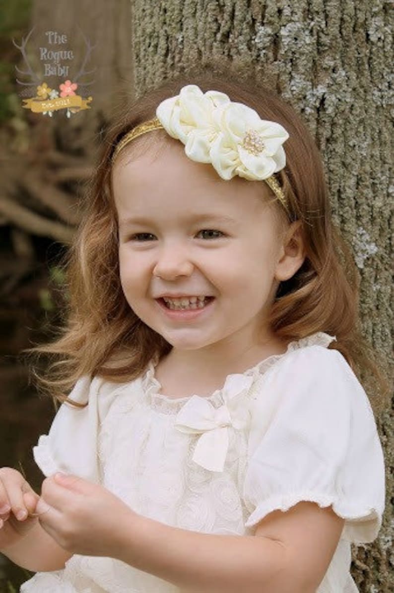 May include: A young girl with long brown hair is wearing a white dress with a lace overlay and a gold headband with a white flower accent. The headband has a small rhinestone in the centre of the flower.