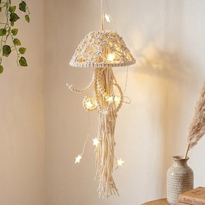 May include: A cream-colored macrame pendant light with a star-shaped string light. The light has a woven lampshade and long, dangling fringe. The light hangs from a wooden beam. A small wooden table holds books, a candle, and a vase.