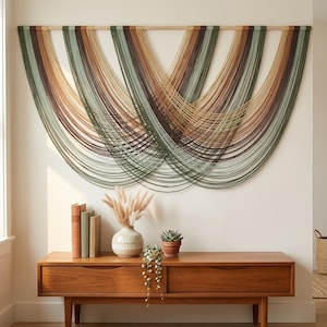 May include: A large wall hanging with a wooden rod and cascading strands of yarn in shades of green, brown, and tan. The artwork is displayed above a wooden console table with a vase, books, and plants, creating a modern, minimalist aesthetic.