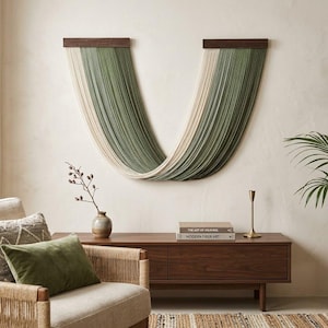 May include: A large wall hanging featuring a U-shaped design made of green, off-white, and gray yarn. The yarn is attached to two dark brown wooden bars. The wall art is displayed above a wooden cabinet in a living room setting.