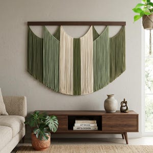 May include: A wall hanging featuring long, vertical strands of sage green and cream-colored cord suspended from a dark brown wooden bar. The cords are arranged in a layered, scalloped pattern, creating a wide, curved shape against the wall.