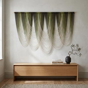 Op de afbeelding: Decoratieve wandophanging met groene, zwarte en witte strengen, opgehangen aan een houten staaf. Het kunstwerk hangt boven een houten kast. Een vaas en boeken staan op de kast.
