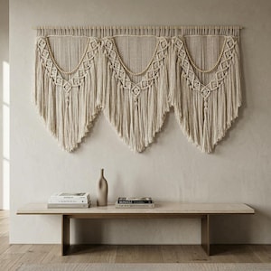 May include: A large, cream-colored macrame wall hanging with a geometric design and beaded accents. The wall decor features a series of triangular shapes with long fringe. Below, a light-colored bench holds books and a vase.