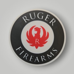 Op de afbeelding: Cirkelvormig embleem met de woorden "RUGER FIREARMS" in witte letters langs de buitenrand. Het midden toont een rood adelaarsymbool met de letter "R" in het midden, tegen een witte achtergrond, met een zwarte rand.