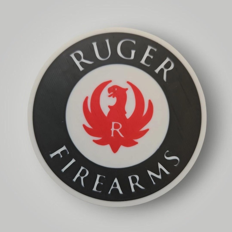 Op de afbeelding: Cirkelvormig embleem met de woorden "RUGER FIREARMS" in witte letters langs de buitenrand. Het midden toont een rood adelaarsymbool met de letter "R" in het midden, tegen een witte achtergrond, met een zwarte rand.
