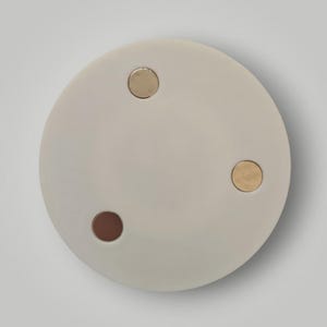 Op de afbeelding: Een rond, lichtgrijs decoratief object met drie cirkelvormige inzetstukken. Twee inzetstukken zijn goudkleurig en &eacute;&eacute;n is donkerbruin. Het object staat tegen een witte achtergrond.