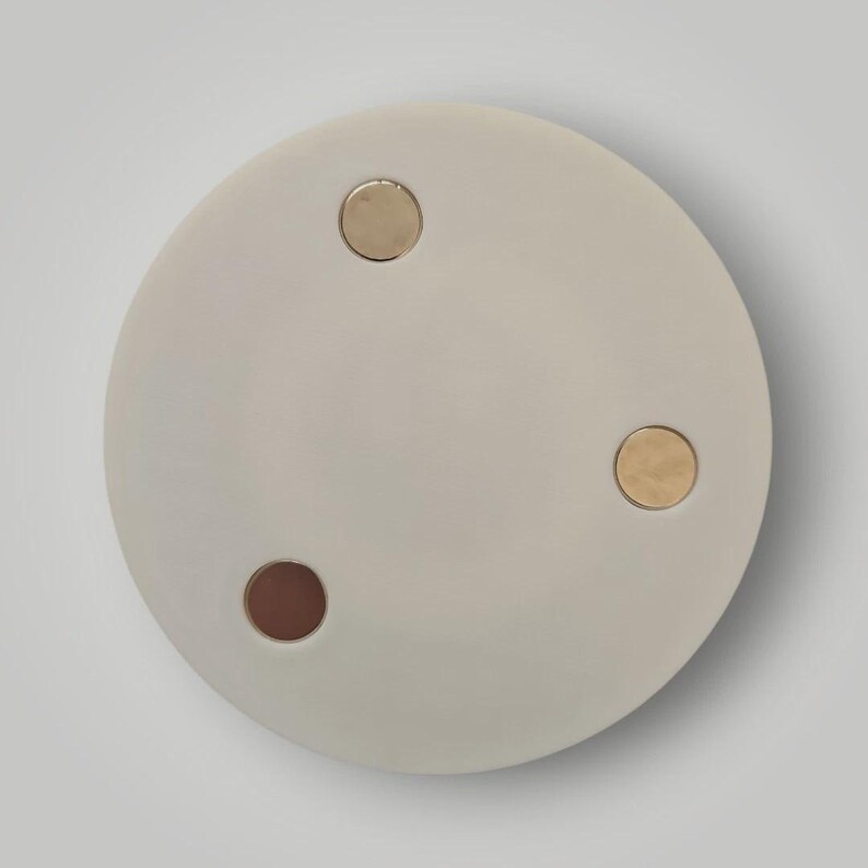 Op de afbeelding: Een rond, lichtgrijs decoratief object met drie cirkelvormige inzetstukken. Twee inzetstukken zijn goudkleurig en &eacute;&eacute;n is donkerbruin. Het object staat tegen een witte achtergrond.