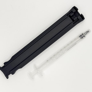 May include: A black, foldable, rectangular object and a clear syringe with measurement markings are displayed on a white surface. The black object appears to be a stand or holder. The syringe has a plunger and needle.