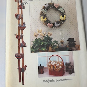 May include: A vintage Simplicity pattern featuring various country-style crafts. The image showcases a wooden wall hanging with fabric chickens and hearts, a wreath with heart and bird accents, and a basket filled with eggs. The pattern is by Marjorie Puckett.