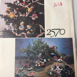 Puede incluir: Vogue Patterns 2570. La imagen muestra una portada de patrón vintage con un árbol de Navidad decorado con adornos hechos a mano y una escena del bosque con figuras de animales. La portada es de color crema con el logotipo de Vogue Patterns.