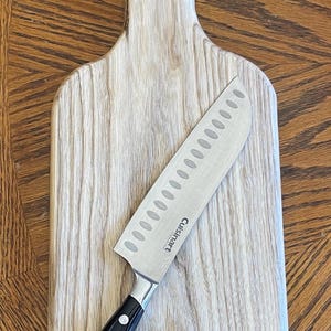 May include: A light-colored wooden cutting board with a stainless steel knife. The knife has a black handle and the brand name "Cuisinart" is visible on the blade. The cutting board is rectangular with a rounded handle.