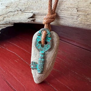 Op de afbeelding: Een unieke hanger met een verweerde, turquoise sleutel op een gladde, grijze steen, opgehangen aan een bruin, gevlochten koord. De sleutel heeft gedetailleerde patronen en is op de steen gemonteerd, wat een rustieke, vintage esthetiek creëert. De steen is ongeveer 5 cm lang.