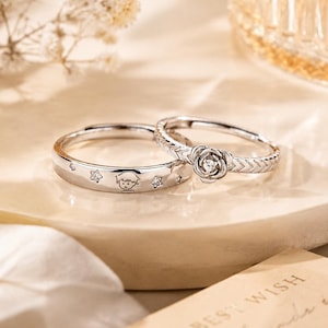 Könnte beinhalten: Zwei silberne Ringe. Ein Ring zeigt einen kleinen Cartoon-Charakter und Sterne. Der andere Ring hat ein Rosenmuster mit einem kleinen Edelstein in der Mitte. Die Ringe liegen auf einer weißen Oberfläche mit unscharfem Hintergrund.