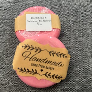Puede incluir: Dos pastillas de jabón redondas de color rosa envueltas en plástico transparente. La pastilla superior tiene una etiqueta de papel color canela que dice Revitalizing & Balancing for Normal Skin. La inferior tiene una etiqueta Handmade come from nature.