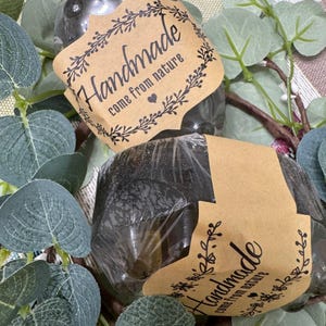 May include: Two dark, translucent, rounded bars of soap wrapped in clear plastic. Each bar features a tan paper band printed with the text "Handmade come from nature" in black ink, decorated with a small heart and leafy vine illustrations.