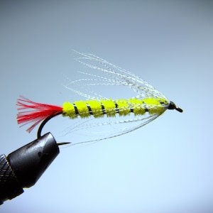 May include: A close-up of a handmade fishing fly. The fly features a yellow body with black stripes, a red tail, and white feather wings. The fly is attached to a black hook.