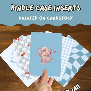 May include: A set of rectangular Kindle case inserts printed on cardstock. The designs include a light blue background with a central illustration of a winged rabbit holding a bow and arrow, a blue and white checkered pattern with red cherries, and a blue wavy checkerboard pattern. The text reads KINDLE CASE INSERTS, PRINTED ON CARDSTOCK, and KAWAII.