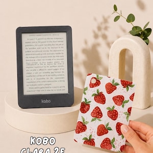 May include: A black Kobo Clara 2E e-reader displaying text on its screen, positioned next to a rectangular protective cover featuring a repeating pattern of red strawberries with green leaves on a white background. The text Kobo Clara 2E is visible.