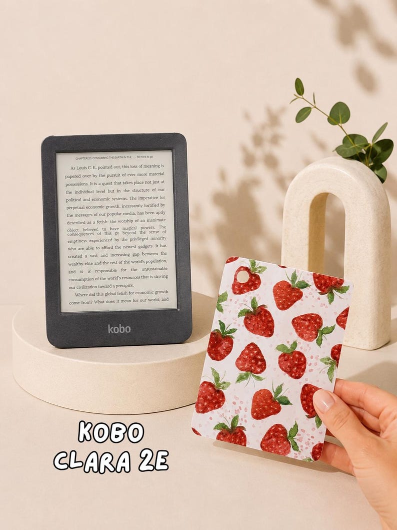 May include: A black Kobo Clara 2E e-reader displaying text on its screen, positioned next to a rectangular protective cover featuring a repeating pattern of red strawberries with green leaves on a white background. The text Kobo Clara 2E is visible.