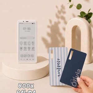 May include: A white Boox Palma e-reader displaying a digital interface with the time 07:00, alongside two rectangular protective cases: one with light blue vertical stripes and the text to live for the hope of it all, and one solid navy blue with the name Olivia.