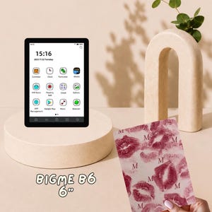 May include: A Bigme B6 6-inch e-reader with a black frame displaying a digital interface with app icons and time. Beside it is a rectangular cover featuring a repeating pattern of dark pink lipstick kiss marks and the letter M on a light background.