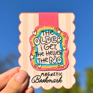 Può includere: Un segnalibro magnetico rettangolare con bordi smerlati e uno sfondo a righe color pesca chiaro. Il centro presenta una grafica con bordo arcobaleno con il testo "THE OLDER I GET, THE HELLER THE NO" e "MAGNETIC Bookmark" in basso.