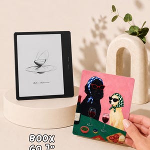 May include: A black Boox Go 7 inch e-reader displaying a sketch of a bowl on its screen, positioned next to a rectangular print featuring a black dog and a light-colored dog wearing headscarves and sunglasses while sitting at a table with wine.