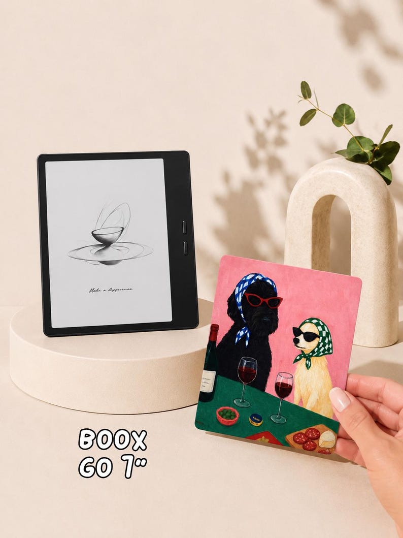 May include: A black Boox Go 7 inch e-reader displaying a sketch of a bowl on its screen, positioned next to a rectangular print featuring a black dog and a light-colored dog wearing headscarves and sunglasses while sitting at a table with wine.