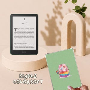 May include: A black Kindle Colorsoft e-reader displaying text on its screen, positioned next to a light green protective case featuring a graphic of a tea bag with the text LGBTea and a small rainbow flag tag, held by a hand.