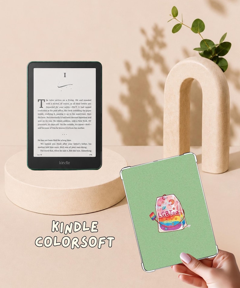 May include: A black Kindle Colorsoft e-reader displaying text on its screen, positioned next to a light green protective case featuring a graphic of a tea bag with the text LGBTea and a small rainbow flag tag, held by a hand.