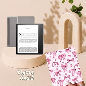 May include: A Kindle Oasis e-reader with a black frame and a screen displaying text titled Book One. Beside it is a rectangular cover featuring a repeating pattern of pink bows and small hearts on a white background with thin vertical pink stripes.