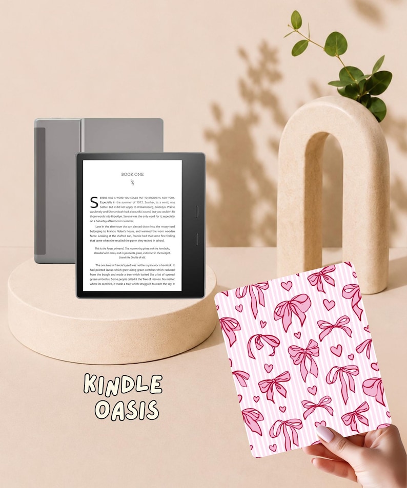 May include: A Kindle Oasis e-reader with a black frame and a screen displaying text titled Book One. Beside it is a rectangular cover featuring a repeating pattern of pink bows and small hearts on a white background with thin vertical pink stripes.