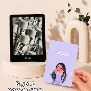 May include: A black Kindle Paperwhite e-reader displaying a black and white image of pencil erasers on its screen. Next to it is a rectangular light purple sticker featuring a cartoon illustration of a person and the text MEGHAN'S KINDLE.