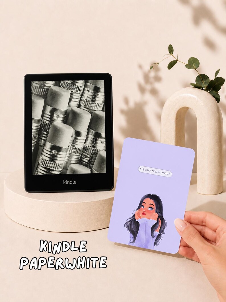 May include: A black Kindle Paperwhite e-reader displaying a black and white image of pencil erasers on its screen. Next to it is a rectangular light purple sticker featuring a cartoon illustration of a person and the text MEGHAN'S KINDLE.