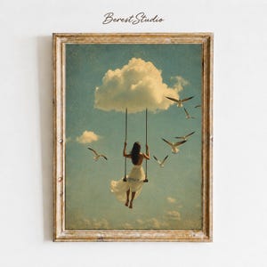 May include: A framed wall art print featuring a person in a white dress sitting on a swing suspended in a blue sky with clouds and flying seagulls. The text Berest Studio is printed at the top of the artwork, which has a vintage texture.
