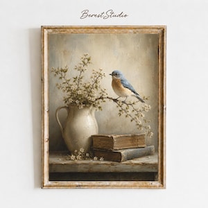 May include: A framed wall art print featuring a blue bird perched on a flowering branch extending from a cream-coloured ceramic pitcher. Two weathered, stacked books sit on a wooden surface below the pitcher. The text Berest Studio is visible.