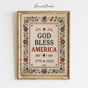 Op de afbeelding: Een ingelijst kruissteek borduurwerk op crèmekleurige stof met een bloemenrand in rood, blauw en groen. De tekst luidt GOD BLESS AMERICA in blauw en rood, met de jaartallen 1776 en 2026 gescheiden door een rode ster, in een houten lijst.
