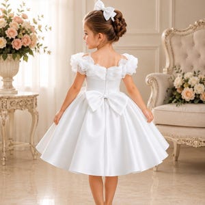 May include: A young girl in a white satin dress with a large bow on the back and a matching bow in her hair. The dress has ruffled sleeves and a full skirt. She is wearing white shoes. The background includes a floral arrangement and a decorative chair.