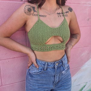 May include: A green crochet halter top with a keyhole design. The top is made of a textured, open-weave fabric. The model is wearing blue jeans. The background is a pink brick wall.