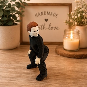 May include: A small figurine of a character with a white mask, brown hair, and a black jumpsuit, posed in a crouching position. The figure has pale skin, dark eye sockets, and is wearing black shoes, capturing a detailed representation of a horror movie icon.