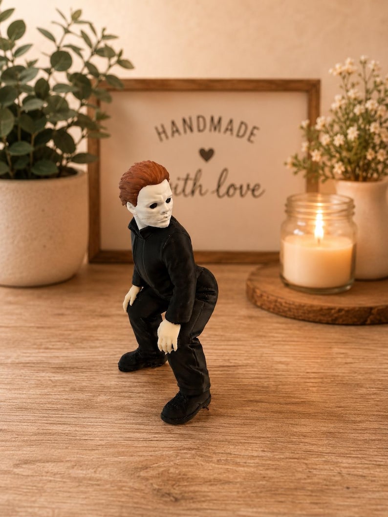 May include: A small figurine of a character with a white mask, brown hair, and a black jumpsuit, posed in a crouching position. The figure has pale skin, dark eye sockets, and is wearing black shoes, capturing a detailed representation of a horror movie icon.